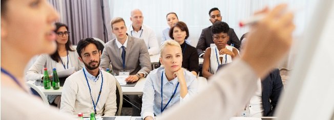 Group of people sitting in a classroom environment watching a woman write on a whiteboard